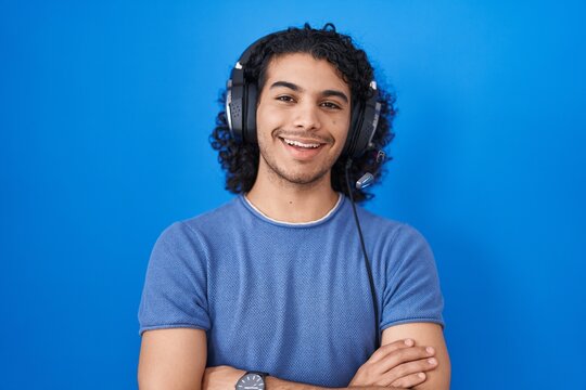 Hispanic man with curly hair listening to music using headphones happy face smiling with crossed arms looking at the camera. positive person.