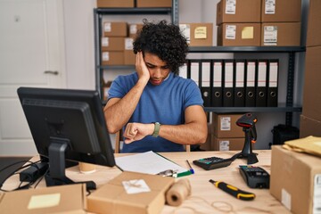 Hispanic man with curly hair working at small business ecommerce looking at the watch time worried, afraid of getting late