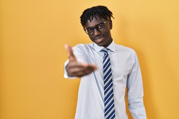 African man with dreadlocks standing over yellow background smiling friendly offering handshake as greeting and welcoming. successful business.