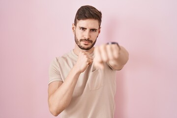 Hispanic man with beard standing over pink background punching fist to fight, aggressive and angry...