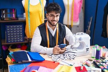 Young hispanic man tailor smiling confident using smartphone at sewing studio