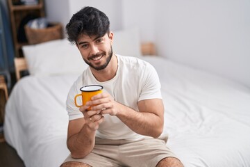 Young hispanic man drinking cup of coffee sitting on bed at bedroom