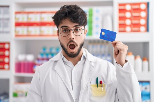 Hispanic man with beard working at pharmacy drugstore holding credit card scared and amazed with open mouth for surprise, disbelief face