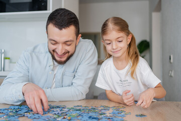 Fototapeta premium Father and daughter playing puzzles at home. Family connecting jigsaw puzzle pieces in a kitchen table. Kid assembling a jigsaw puzzle. Fun family leisure. Stay at home activity for kids.