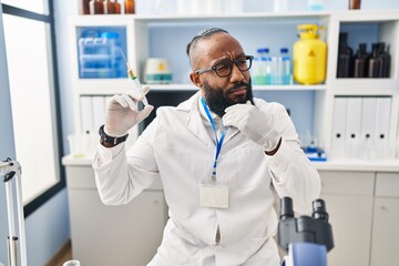 Naklejka na ściany i meble African american man working at scientist laboratory holding syringe serious face thinking about question with hand on chin, thoughtful about confusing idea