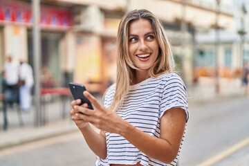 Young blonde woman smiling confident using smartphone at street