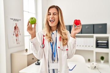 Young blonde doctor woman holding heart and green apple winking looking at the camera with sexy...