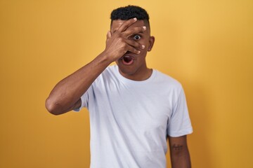 Young hispanic man standing over yellow background peeking in shock covering face and eyes with hand, looking through fingers with embarrassed expression.