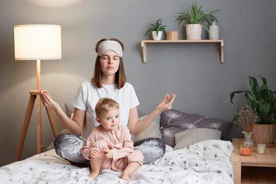 Image of exhausted mother and infant kid at home, woman sitting in lotus pose and practicing yoga, trying to relax and calm down, keeps eyes closed, baby daughter playing near mom.