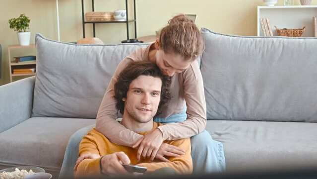 Medium shot of affectionate young Caucasian couple sitting together in living room watching TV on weekend