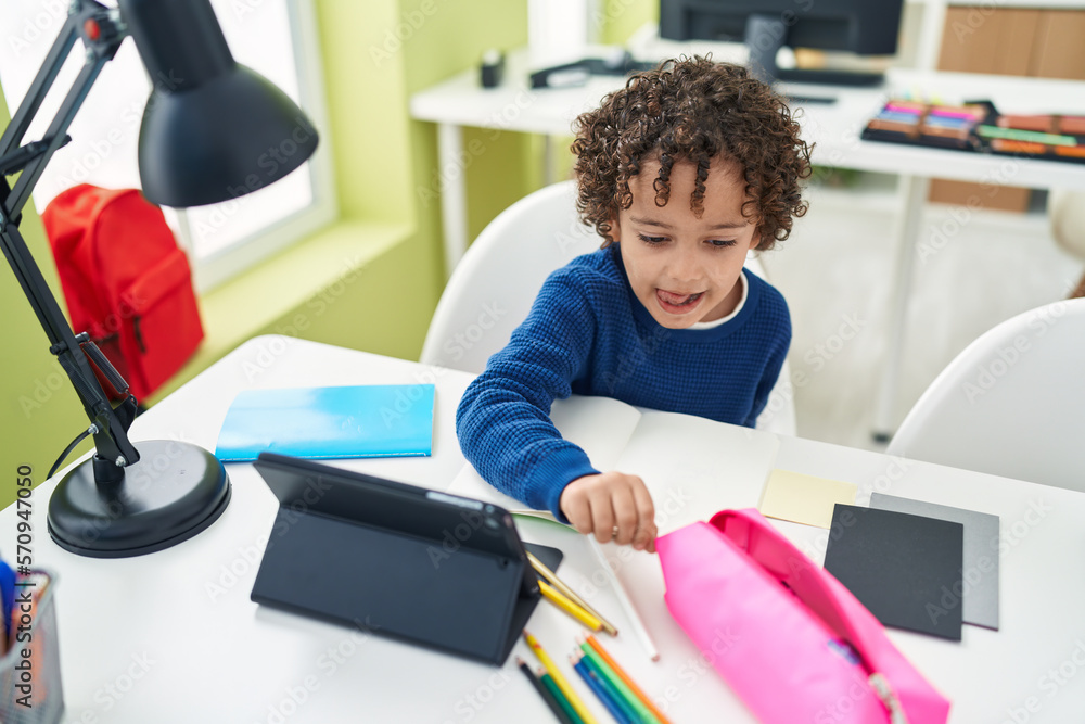 Sticker Adorable hispanic boy preschool student sitting on table drawing on notebook at classroom