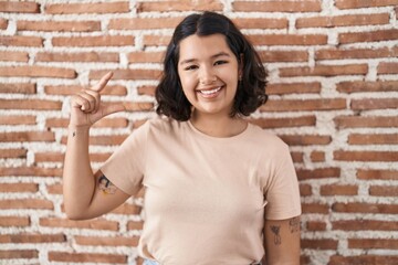 Young hispanic woman standing over bricks wall smiling and confident gesturing with hand doing small size sign with fingers looking and the camera. measure concept.