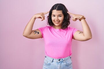 Obraz na płótnie Canvas Young hispanic woman standing over pink background smiling pointing to head with both hands finger, great idea or thought, good memory