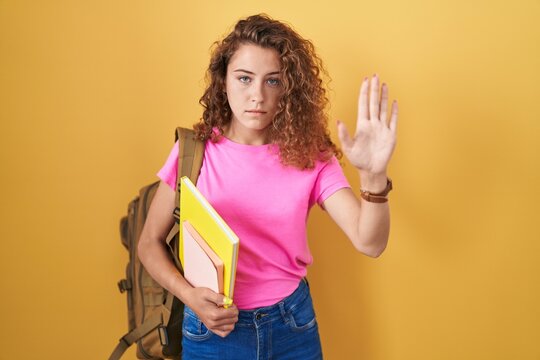 Young caucasian woman wearing student backpack and holding books doing stop sing with palm of the hand. warning expression with negative and serious gesture on the face.