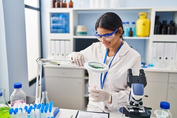 Young latin woman wearing scientist uniform using magnifying glass at laboratory