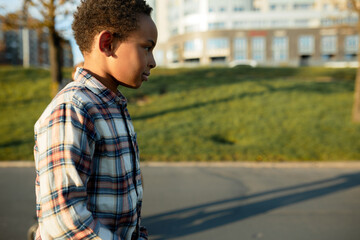 Profile view of handsome african american kid boy returning home from walk, going down city streets on sidewalk leaving shadow on sun-drenched concrete with blurred background of multistory buildings