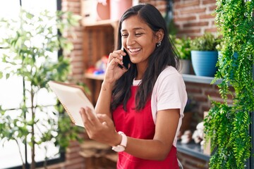 Young beautiful hispanic woman florist talking on smartphone reading notebook at flower shop