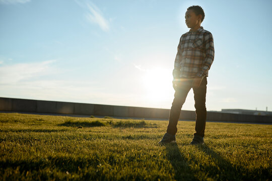 Outdoor image of african american male kid in casual clothes standing in middle of lawn in rays of evening sun during his walk in public city park on weekend, looking aside on copy space