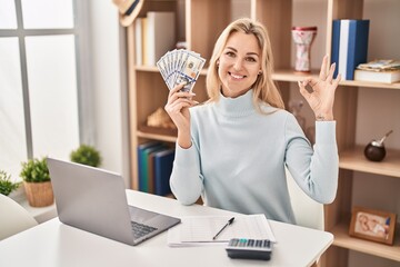 Young caucasian woman using laptop holding dollars banknotes doing ok sign with fingers, smiling friendly gesturing excellent symbol