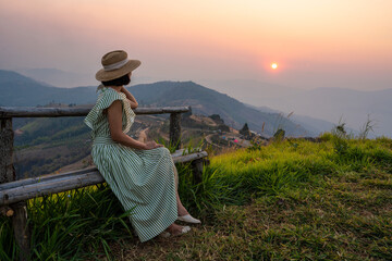 Asian tourist girl in dress wearing hat sit and relax watching mountain view during sunset tourist destination trekking to see nature during summer vacation