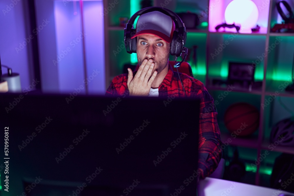 Poster young caucasian man playing video games covering mouth with hand, shocked and afraid for mistake. su