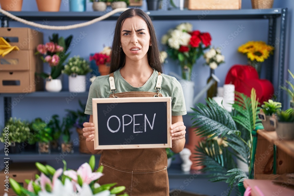 Wall mural Young brunette woman working at florist holding open sign clueless and confused expression. doubt concept.