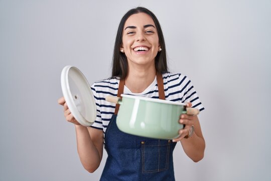 Young brunette woman wearing apron holding cooking pot smiling and laughing hard out loud because funny crazy joke.