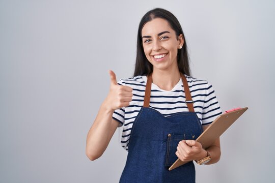 Young brunette woman wearing professional waitress apron and clipboard smiling happy and positive, thumb up doing excellent and approval sign