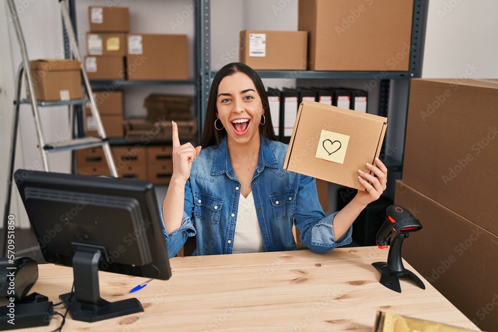 Canvas Prints Hispanic woman working at small business ecommerce holding box surprised with an idea or question pointing finger with happy face, number one