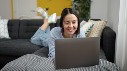 Young beautiful hispanic woman using laptop lying on sofa at home