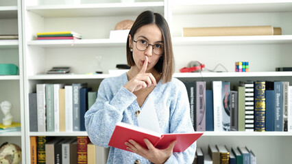 Young beautiful hispanic woman student reading book doing silence gesture at library university