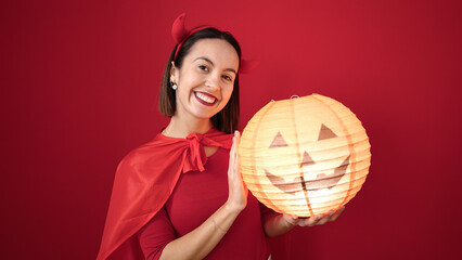 Young beautiful hispanic woman wearing devil costume holding halloween pumpkin lamp over isolated red background