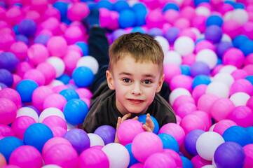 A beautiful, happy, small, smiling boy, a preschool child lies in a variety of multi-colored, colored plastic balls on the playground. Photography, portrait, childhood concept.