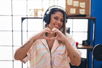 Young hispanic woman musician listening to music doing heart gesture with hands at music studio