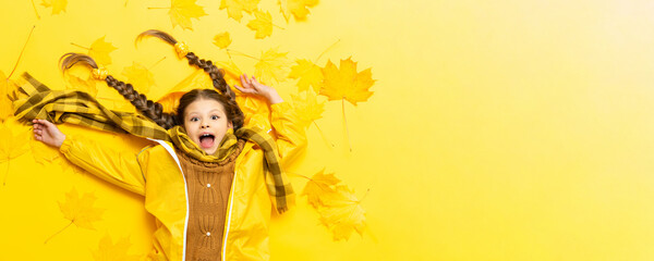 A cheerful little girl in an autumn raincoat and a warm sweater with a scarf on a yellow background...