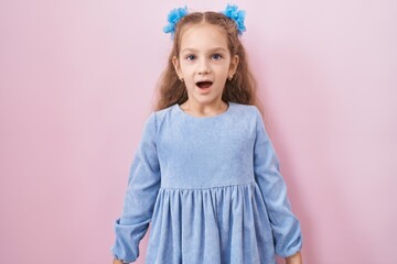 Young little girl standing over pink background scared and amazed with open mouth for surprise,...