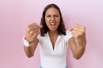 Young hispanic woman wearing casual white t shirt angry and mad raising fists frustrated and...