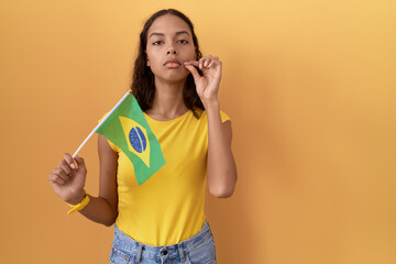 Young hispanic woman holding brazil flag mouth and lips shut as zip with fingers. secret and...