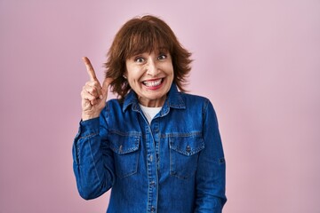 Middle age woman standing over pink background pointing finger up with successful idea. exited and...
