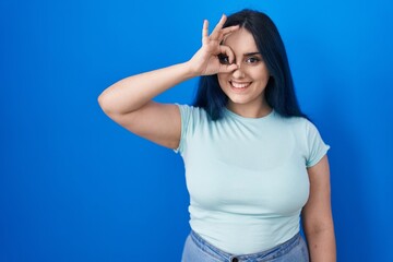 Young modern girl with blue hair standing over blue background doing ok gesture with hand smiling, eye looking through fingers with happy face.