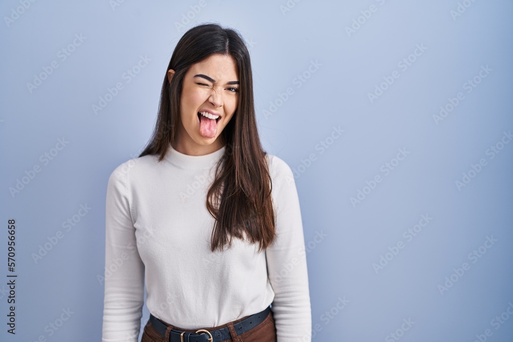 Canvas Prints Young brunette woman standing over blue background sticking tongue out happy with funny expression. emotion concept.