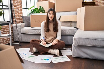 Young brunette woman moving to a new home doing finances scared and amazed with open mouth for...