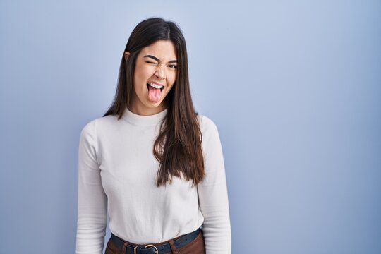 Young brunette woman standing over blue background sticking tongue out happy with funny expression. emotion concept.