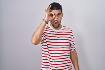 Hispanic man with long hair standing over isolated background doing ok gesture shocked with surprised face, eye looking through fingers. unbelieving expression.