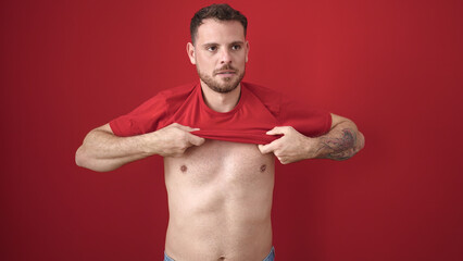 Young caucasian man standing with serious expression taking t shirt off over isolated red background