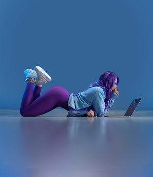 sexy girl in stylish sportswear and with purple hair sexy lying on the floor with a laptop on a blank blue background copy paste