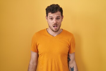 Young hispanic man standing over yellow background in shock face, looking skeptical and sarcastic,...