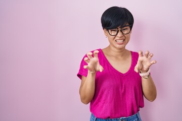 Young asian woman with short hair standing over pink background smiling funny doing claw gesture as...