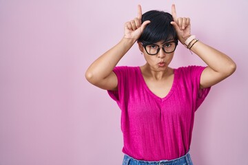 Fototapeta na wymiar Young asian woman with short hair standing over pink background doing funny gesture with finger over head as bull horns