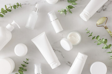 Skincare products concept. Top view photo of white cosmetic bottles, cream jars, dropper bottles, face massage roller and eucalyptus leaves on marble background. Cosmetics mockup idea.